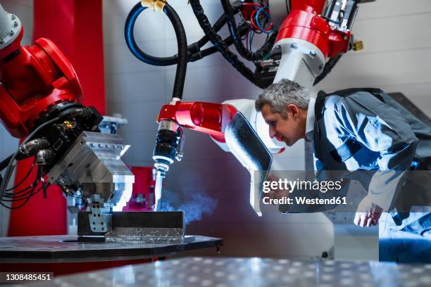 mature worker with welding helmet working at robotics in factory - metallindustrie stock-fotos und bilder