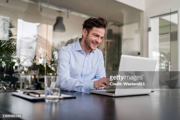 smiling businessman using laptop at desk in office - business man at desk stock-fotos und bilder