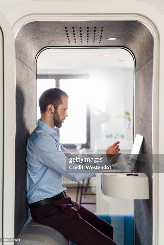 Male entrepreneur attending meeting on laptop in telephone booth at office