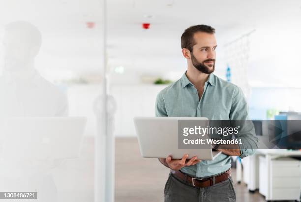 young professional with laptop looking away while standing at office - tre quarti foto e immagini stock