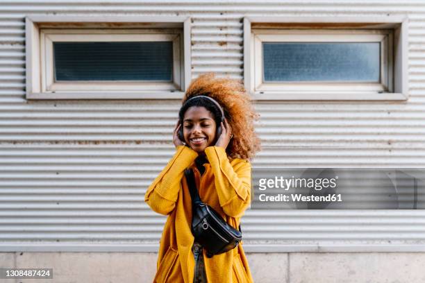 smiling afro woman with eyes closed listening music through wireless headphones while standing against wall - bolsa canguro fotografías e imágenes de stock