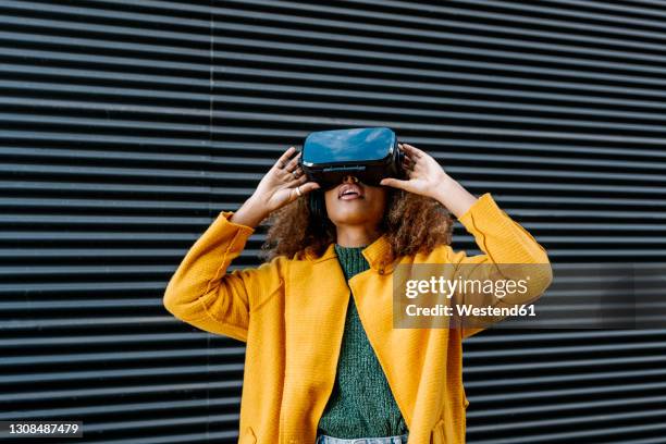 afro woman in warm clothing watching video through virtual reality simulator against wall - réalité virtuelle photos et images de collection