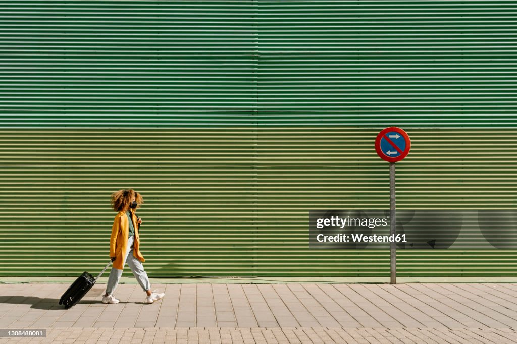 Afro woman with suitcase walking on footpath by green wall during pandemic