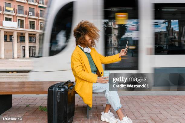 smiling afro woman with tousled hair taking selfie through mobile phone while sitting on bench at tram station - luggage trolley stock-fotos und bilder