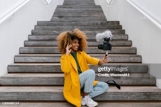 smiling afro woman waving hand during vlogging through camera while sitting on staircase - selfiestick stock-fotos und bilder