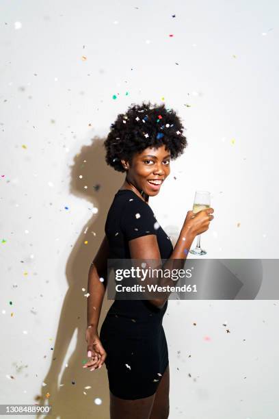 confetti falling on cheerful woman holding champagne flute while standing against white background - woman little black dress stock pictures, royalty-free photos & images