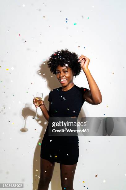 confetti falling on happy woman dancing with champagne flute while standing against white background - fall party inside stock-fotos und bilder