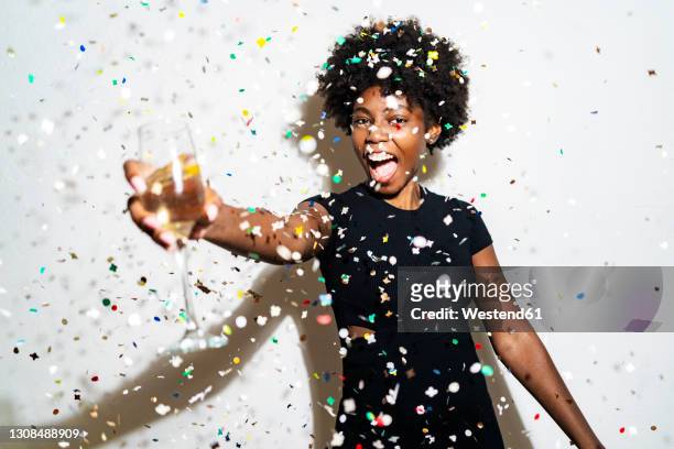 cheerful woman with champagne flute standing amidst confetti against white background - fall party inside stock-fotos und bilder