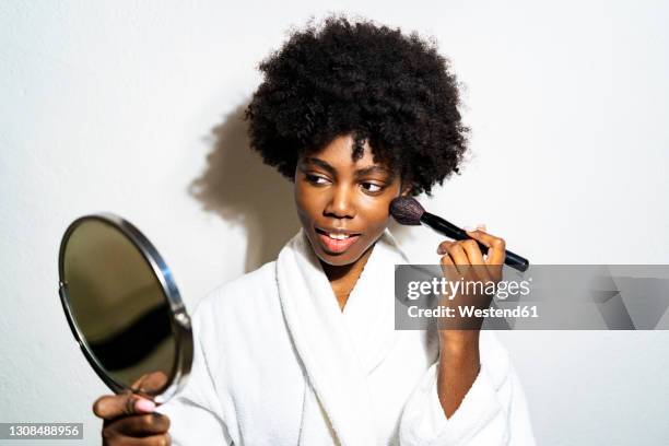 curly hair woman in bathrobe doing make-up while standing against white background - woman applying blush stock pictures, royalty-free photos & images