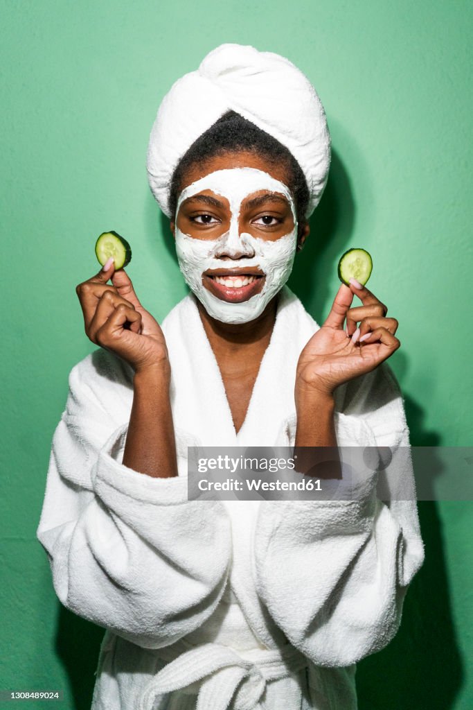 Young woman with facial mask holding cucumber while standing against green background