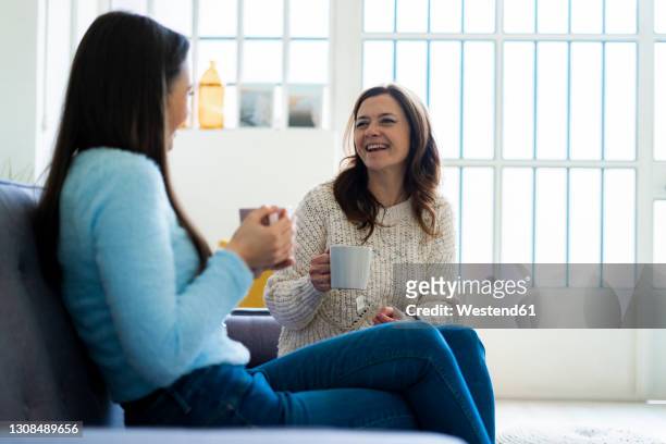 happy mother and daughter with coffee mug sitting on sofa at home - family on couch with mugs stock-fotos und bilder