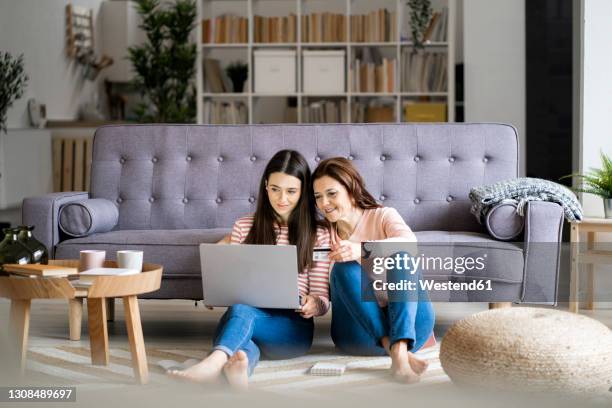 smiling mother holding credit card while daughter paying bills through laptop at home - mother daughter couch imagens e fotografias de stock