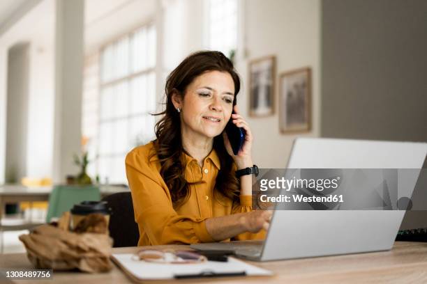 smiling businesswoman using laptop while talking on mobile phone at desk in home office - italian woman stock pictures, royalty-free photos & images