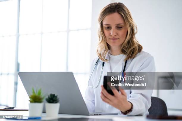 blond female doctor using mobile phone sitting at desk in medical clinic - doctor using smartphone stock-fotos und bilder