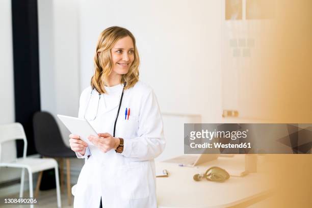 smiling female healthcare worker with tablet leaning on desk while standing in medical clinic - caucasian doctor and nurse using tablet computer stock-fotos und bilder