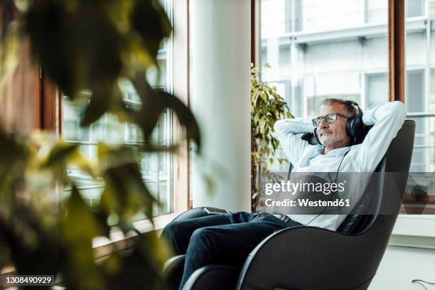 relaxed businessman with hands behind head listening music through headphones while sitting on massage chair in office corridor - massagestuhl stock-fotos und bilder