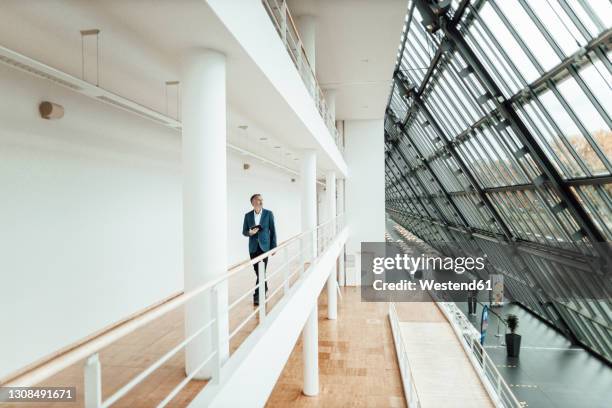 senior male entrepreneur holding digital tablet while walking in corridor at office - distante stock pictures, royalty-free photos & images