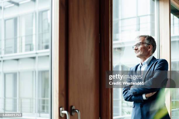 thoughtful man with arms crossed looking through window in office - ビジネスフォーマル ストックフォトと画像