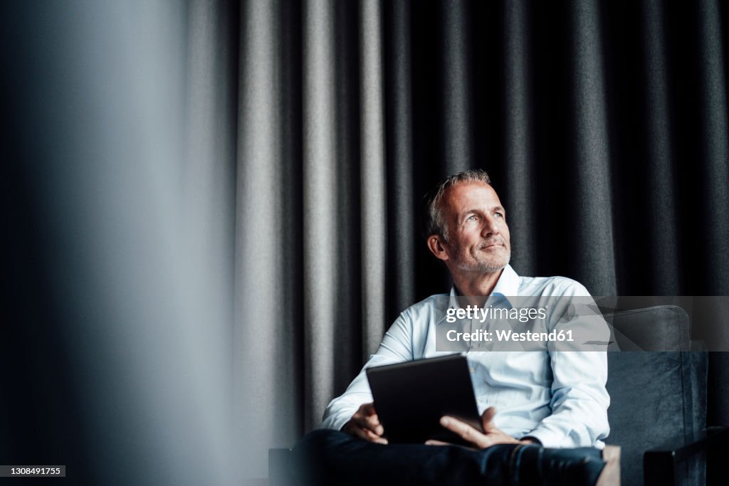 Contemplating businessman with digital tablet looking away while sitting on armchair in office cafeteria