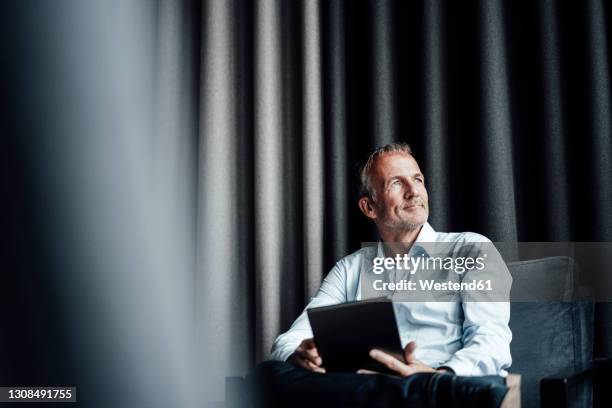 contemplating businessman with digital tablet looking away while sitting on armchair in office cafeteria - casual businessman stockfoto's en -beelden