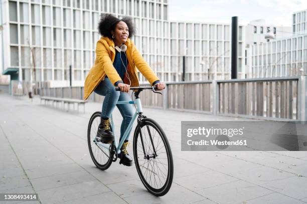 young woman riding bicycle on footpath against building - black woman riding bike stock pictures, royalty-free photos & images