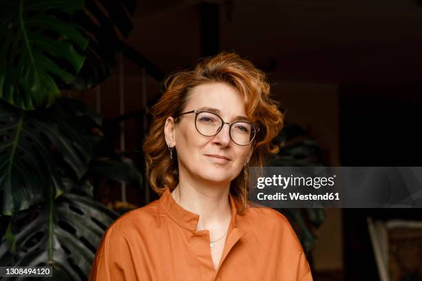 confident mature woman with eyeglasses against plants at home - redheaded women stock-fotos und bilder