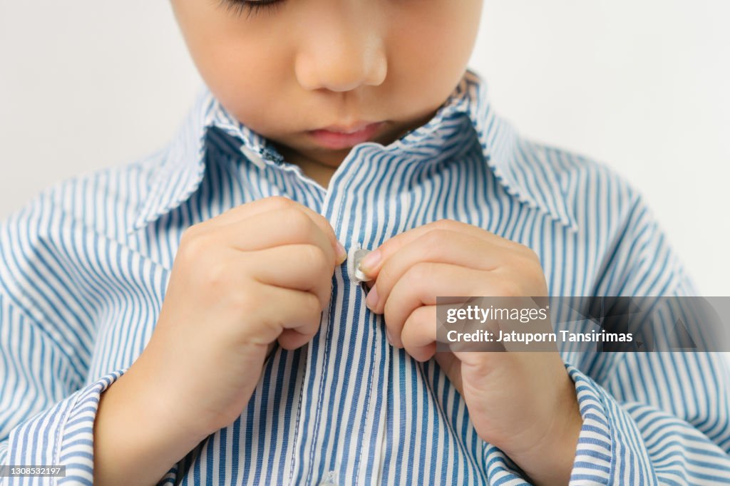 Lindo niño asiático vestido para la escuela, abotonando camisa de manga larga con concentración y felicidad para volver a la escuela