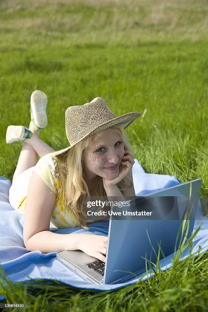 Blond girl wearing a hat, smiling and working on a laptop while lying on a blanket on a meadow