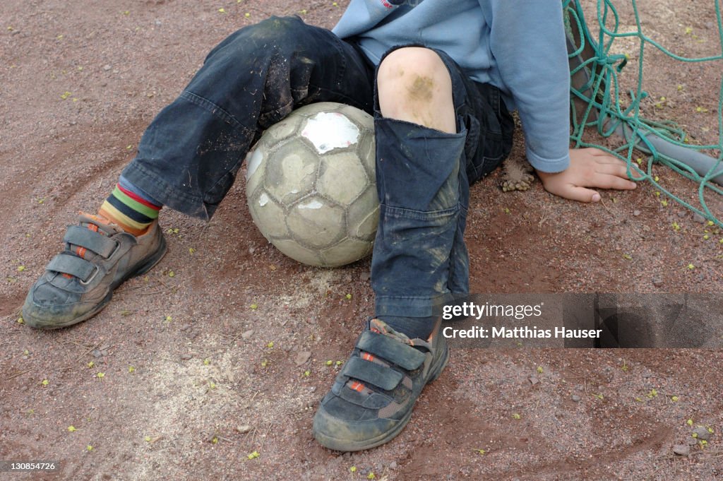 A boy in torn trousers is sitting near the goal with a Football between his knees