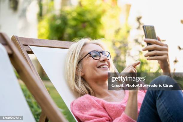 smiling mature woman using smartphone while sitting on deckchair - electronic banking stock pictures, royalty-free photos & images