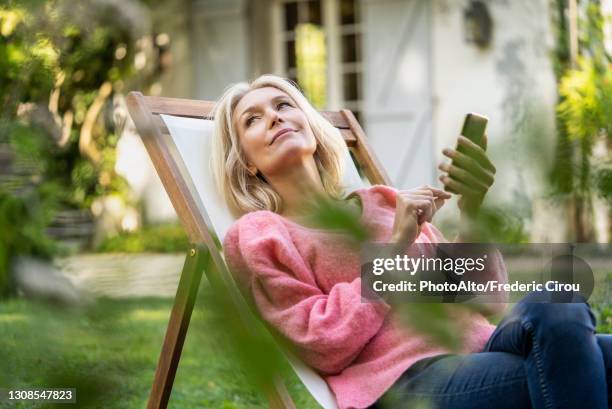 thoughtful mature woman using smartphone while sitting on deckchair - ligstoel stockfoto's en -beelden