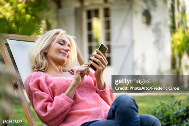 smiling mature woman using smartphone while sitting on deckchair - frau 50 garten stock-fotos und bilder