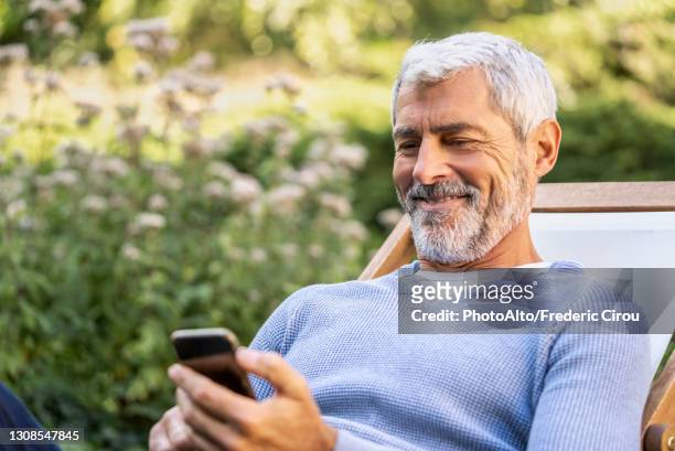 smiling mature man using smartphone while sitting on deckchair - medical insurance stock-fotos und bilder