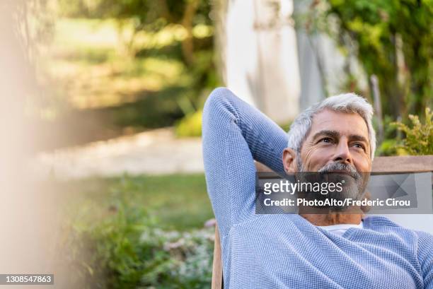 thoughtful mature man with hands behind head sitting on deckchair - alivio fotografías e imágenes de stock