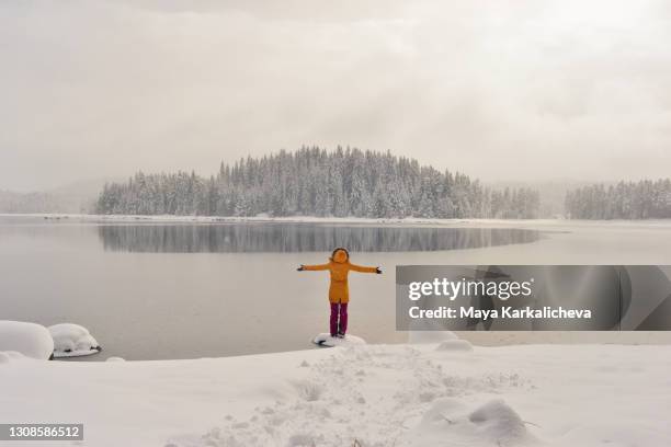 portrait of young woman with arms raised, standing by mountain lake with frozen snowy pine woodland in the background - parka cappotto invernale foto e immagini stock