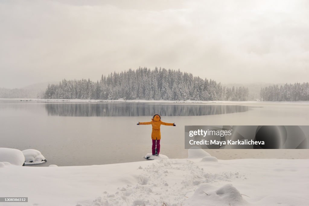 Portrait of young woman with arms raised, standing by mountain lake with frozen snowy pine woodland in the background