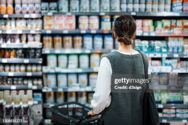 rear view of young asian mother with a shopping cart grocery shopping for baby products in a supermarket. she is standing in front of the baby product aisle and have no idea which product to choose from - precio fotografías e imágenes de stock
