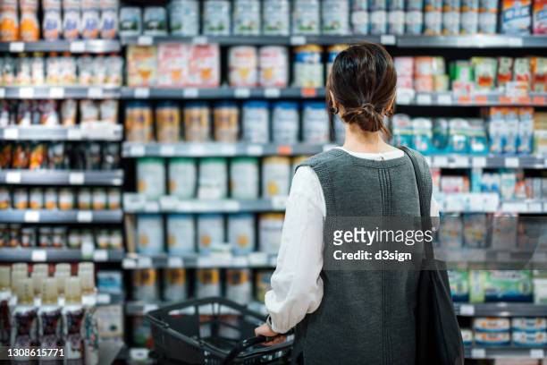 rear view of young asian mother with a shopping cart grocery shopping for baby products in a supermarket. she is standing in front of the baby product aisle and have no idea which product to choose from - faire les courses photos et images de collection