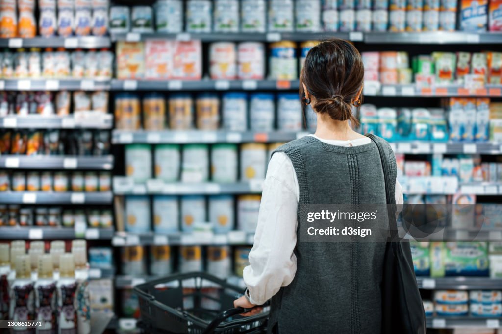 Rear view of young Asian mother with a shopping cart grocery shopping for baby products in a supermarket. She is standing in front of the baby product aisle and have no idea which product to choose from