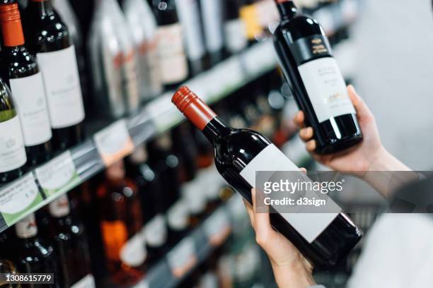 over the shoulder view of woman walking through liquor aisle and choosing bottles of red wine from the shelf in a supermarket - ワインボトル ストックフォトと画像
