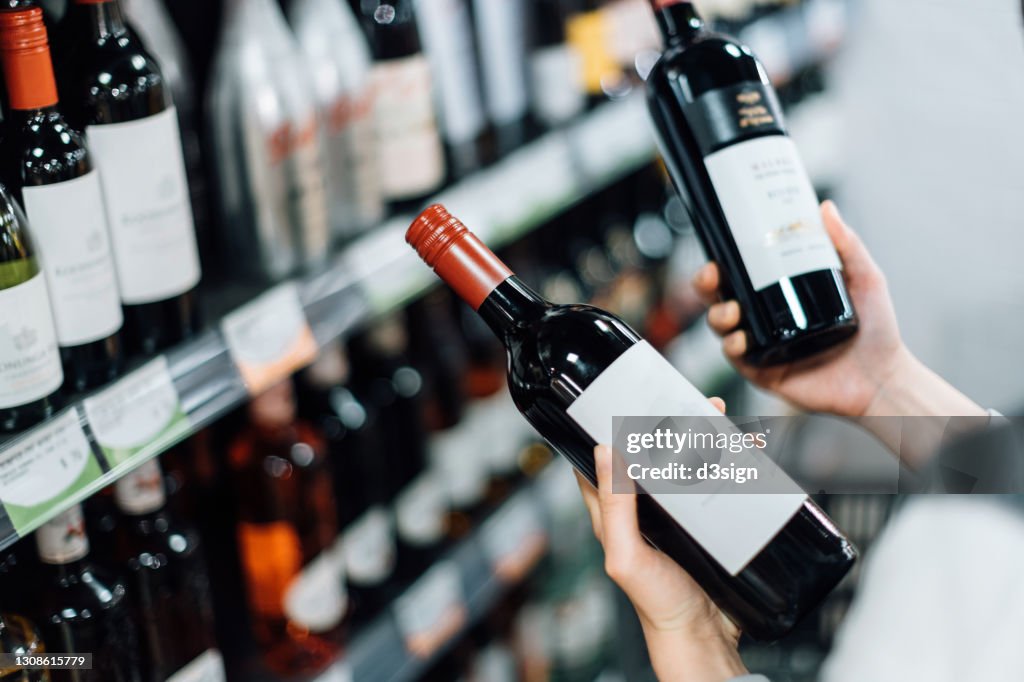Over the shoulder view of woman walking through liquor aisle and choosing bottles of red wine from the shelf in a supermarket