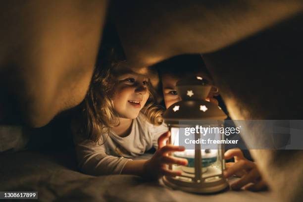 weinig zuster en haar broer die op bed onder deken in donkere ruimte liggen en rustieke lantaarnlamp houden - brother blanket stockfoto's en -beelden