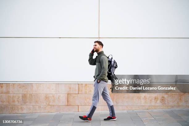 man with backpack talks on phone while walking against white wall - walking side view stock-fotos und bilder