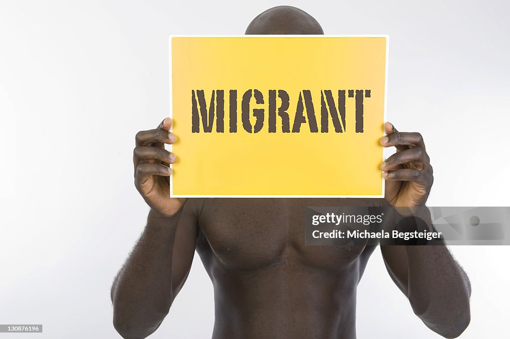Dark-skinned man holding a banner with the word Migrant in front of his face