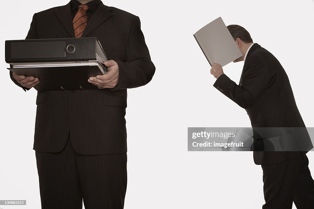 Businessman hiding his face in a file while another businessman walks through the shot, his face cropped