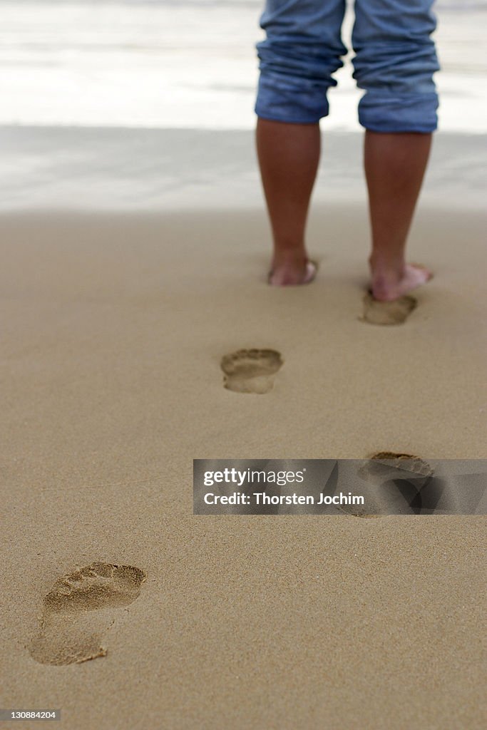 Footprints in the wet sand with legs of an woman in front of the sea.