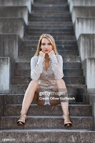 young woman with long blond hair wearing a grey shirt, white shorts and high heels sitting on stone stairs - steintreppe stock-fotos und bilder