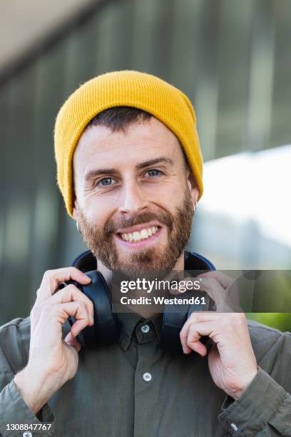 smiling man holding headphones around neck against building - headphone man on neck stock pictures, royalty-free photos & images