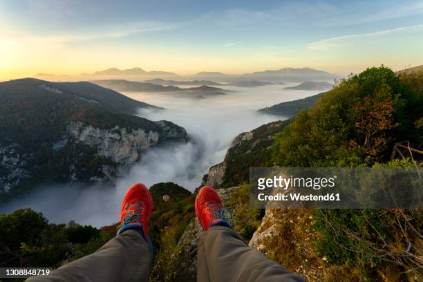 man sitting on mountain against sky during sunrise at furlo gorge, marche, italy - pov shoes stock-fotos und bilder
