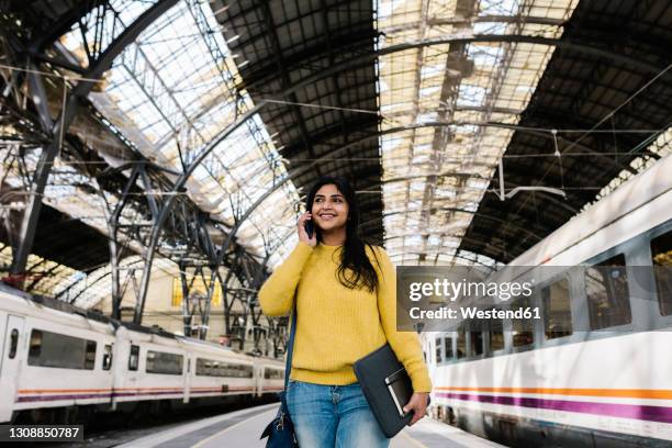 smiling woman with laptop bag looking away while talking on smart phone on railroad station platform - borsa per laptop foto e immagini stock